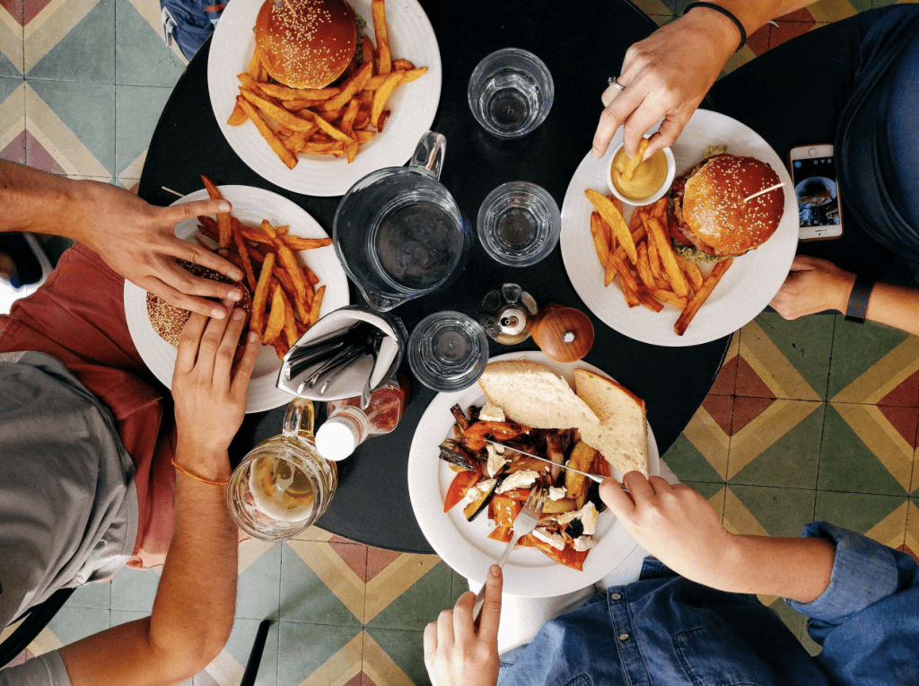 Table setting for four people, eating burgers and sandwiches with fries, tile floor on the background, people holding burgers and fork and knife. 